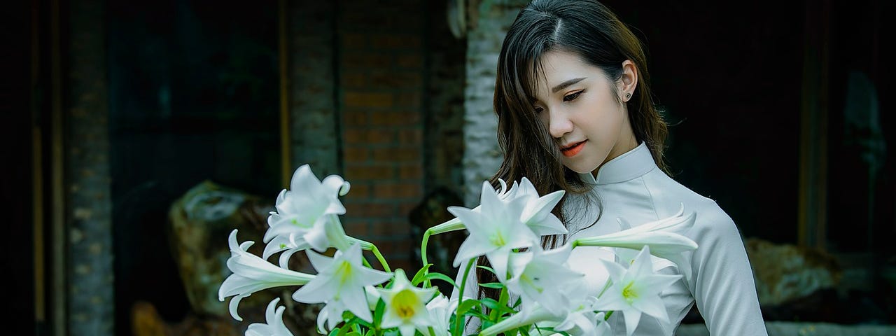 A woman with dark hair wearing white looking down at a large bouquet of white lilies.