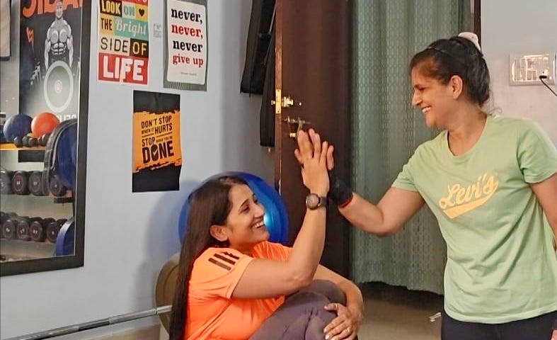 Two women in a gym, one in an orange t-shirt and another in green, high-five each other.