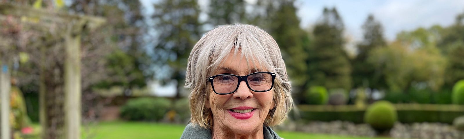 Photograph of actor Sue Johnston stood in her garden. She’s an older woman with short, light-coloured hair and glasses. She’s smiling at the camera.