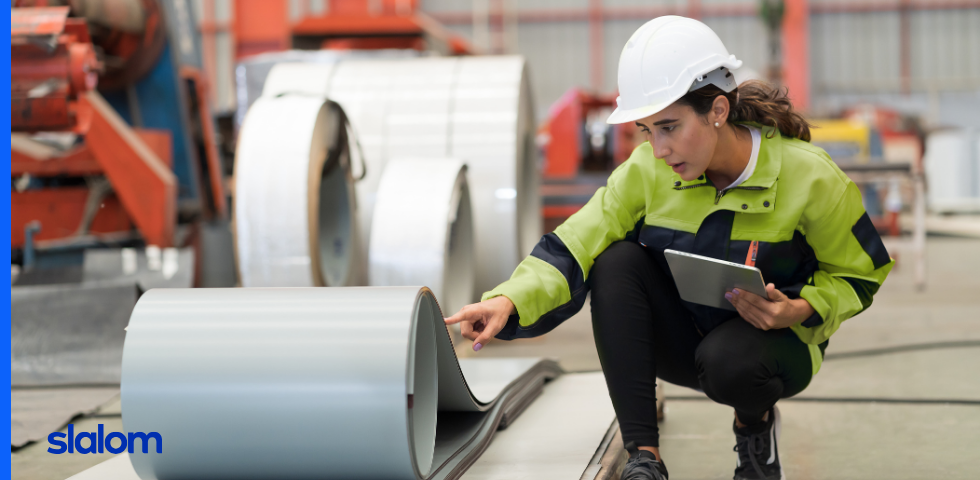 Woman in a hardhat in a warehouse crouching beside a roll of manufactured material