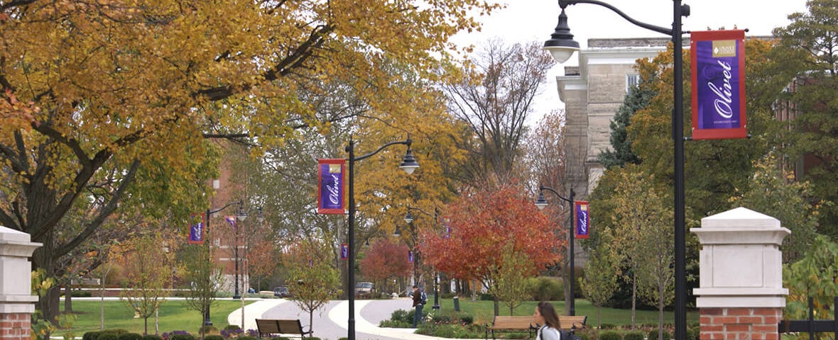 Woman with backpack walking across a campus path at Olivet Nazarene University. There are many trees that line the path and are showing fall colors.