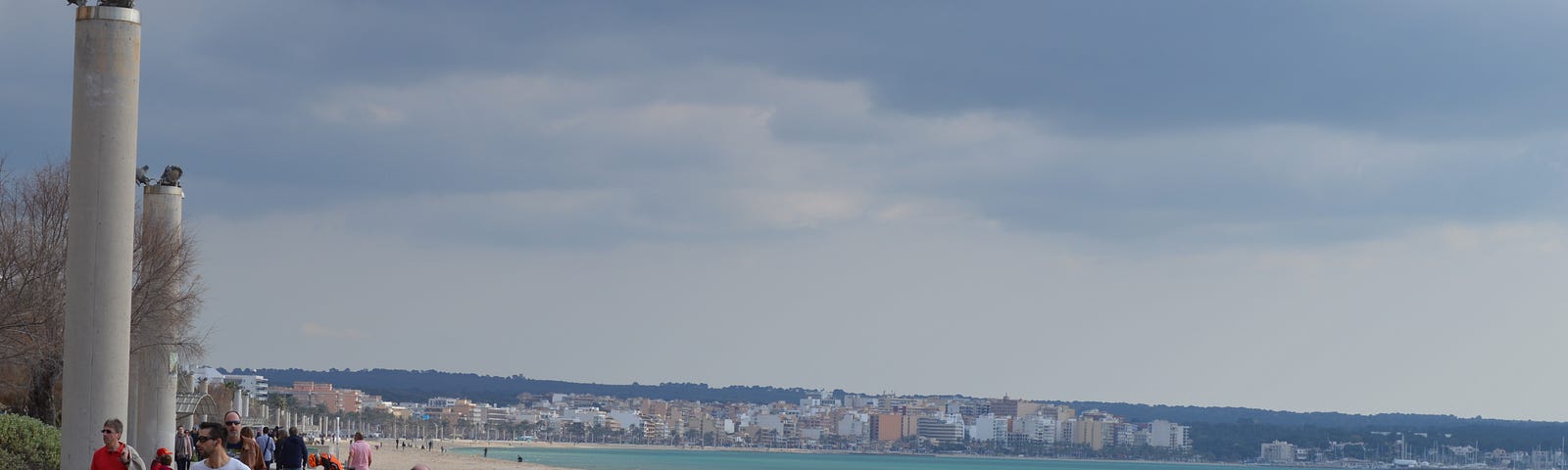 Some people walking on a beach promenade by the sea and some buildings and monuments can be seen in the distance.