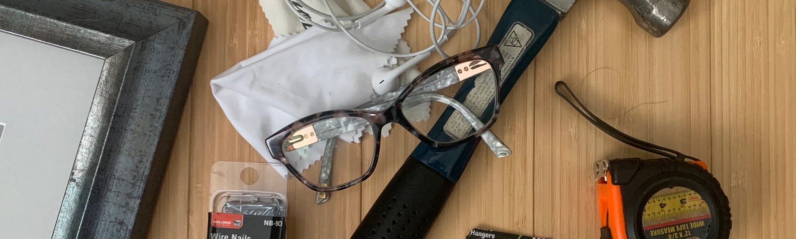 A hammer is surrounded by a measuring tape and variety of picture hangers on a wooden floor. The frame of a painting can also be seen in the frame. On top of the hammer is a pair of bifocals and glasses cleaning cloths, knotted into headphones.