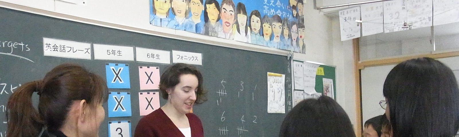 A teacher holds a binder while standing in front of a chalkboard with Japanese writing and Arabic numerals. Five students surround her, engaged with her lesson.