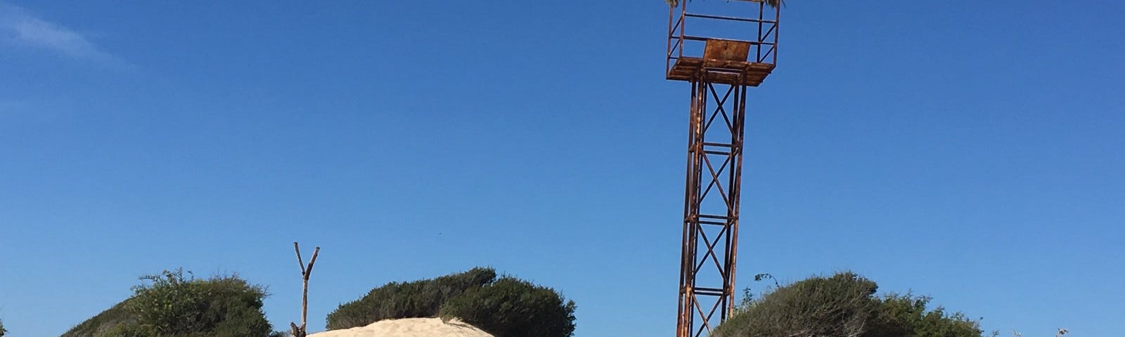 Image of a brilliant blue sky, over a desert sand dune; looking up the sandy dune with green tufts of wild brush to either side and in a couple places at the top. With a clear wide natural path of sand to the top. And a rusted red metal tower of some kind (hurricane watch or other) sticking up into the air at an angle from just over the other side of the dune. The caption reads: “Another hill to climb | from an early walk.” Author’s photo.