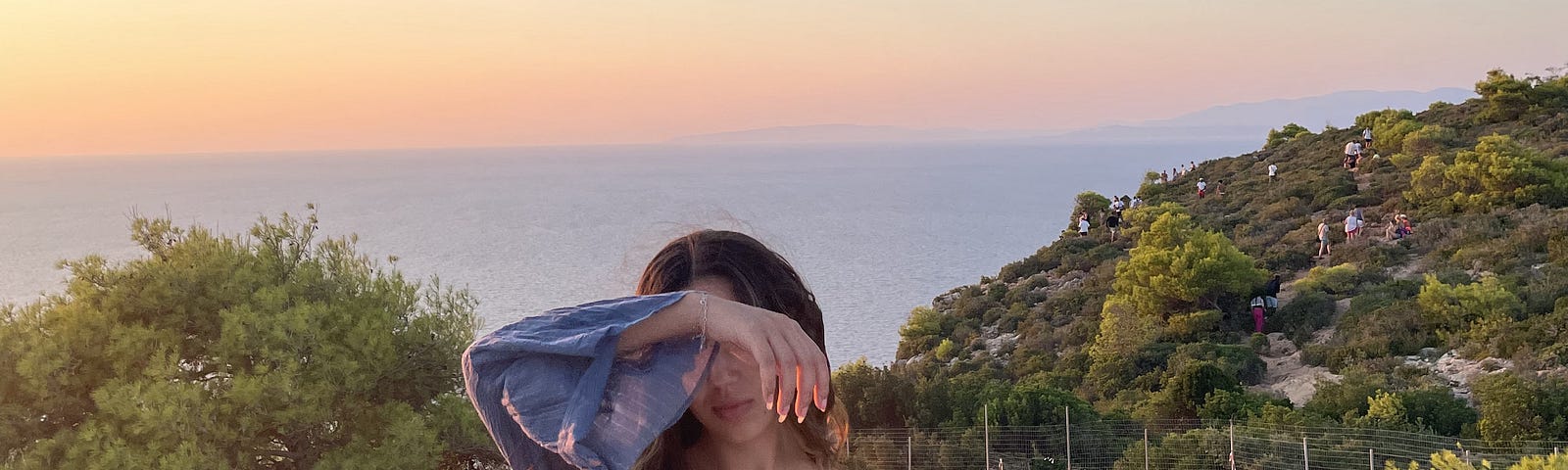 A beautiful landscape in Zakynthos, Greece, with trees, the sea and the sky during a sunset. A girl with long hair is right in the middle wearing blue covering her eyes ina poetic way.