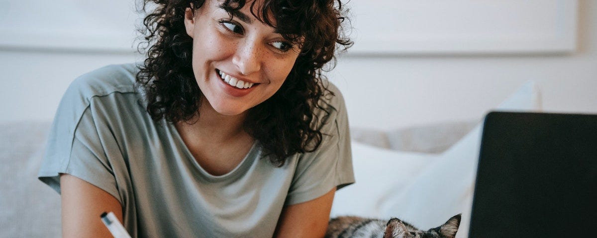 A smiling while looking at her laptop, about to make notes in a notepad. Her cat is staring at the screen in apparent disapproval.