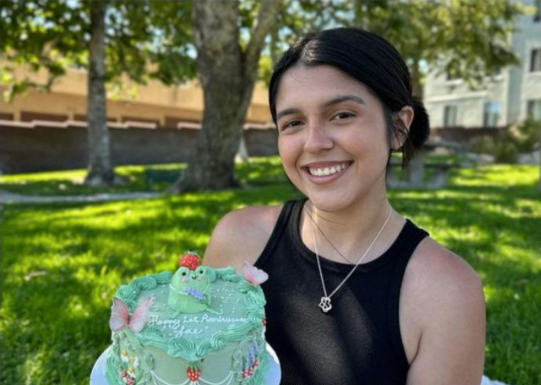 Alissa Portillo holds up a teal cake for the camera. The frame focuses on her head and upper torso, with grass and a few trees filling out the background. Portillo is smiling with her hair in a low bun. She is wearing a black tank top and chain necklace with one charm.