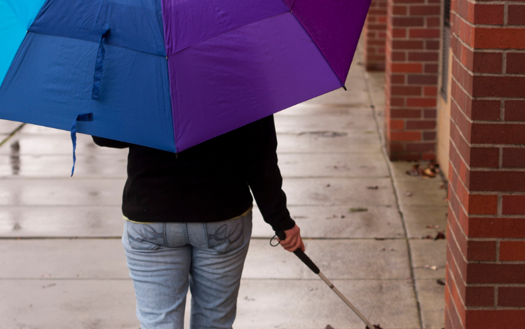 Fotografía de una persona camninando con un bastón blanco en la mano derecha. En la mano izquierda lleva un paraguas con los colores del arcoiris. Crédito: Canva