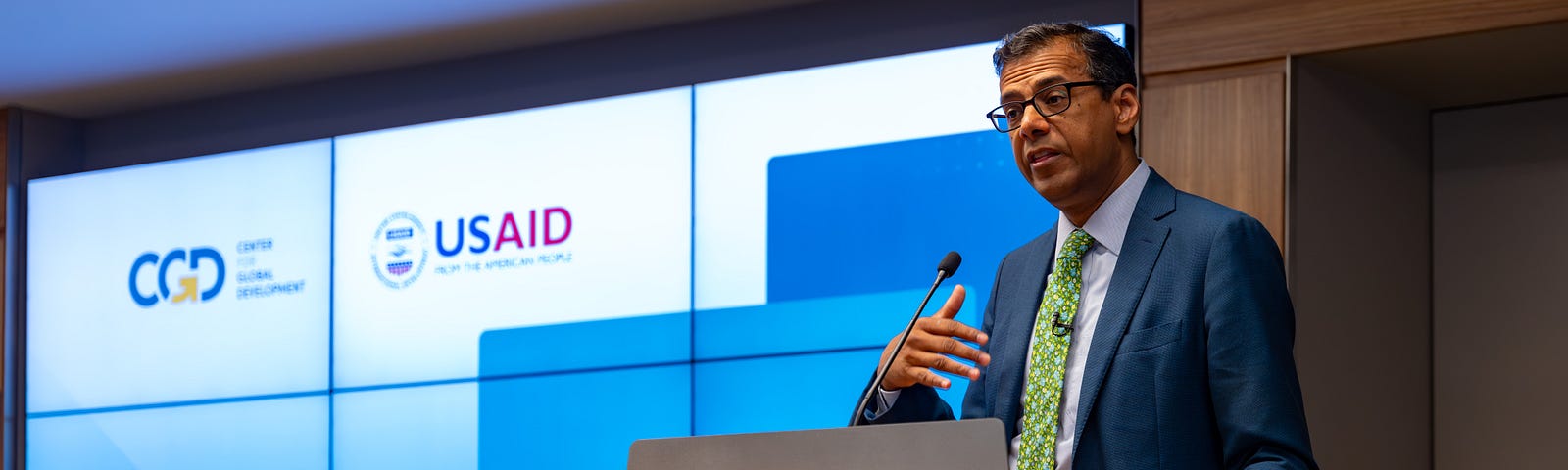 USAID Assistant Administrator for Global Health Dr. Atul Gawande speaks into a microphone while standing behind a podium adorned with the logo CGD. Behind him is an electronic display with the logos of CGD and USAID.
