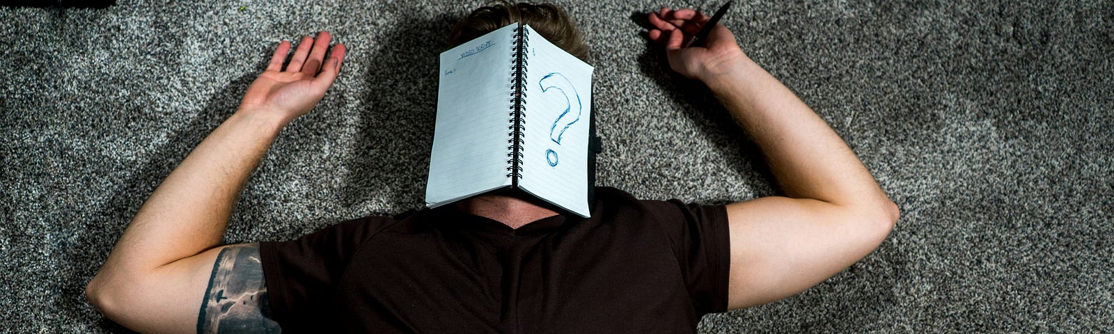 Man in black shirt and black tattoo on inner upper arm lieing on his back on a gray carpet, arms placed by his head with a notebook opened over his face and a big question mark drawn on one page.