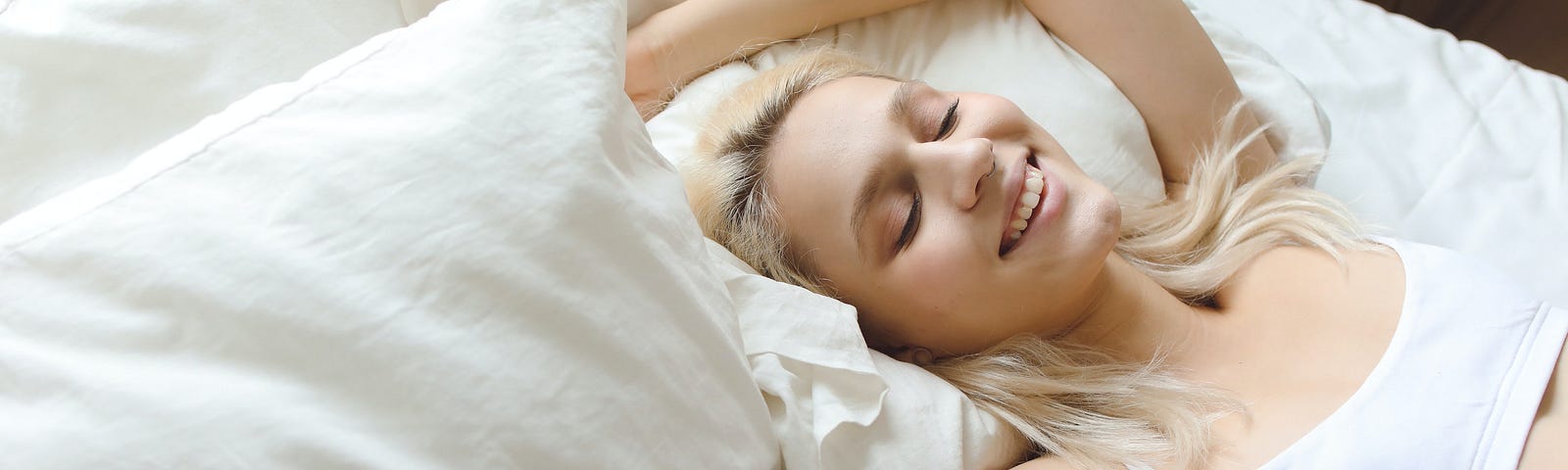 Woman blonde hair white bra top in bed pillows smiling