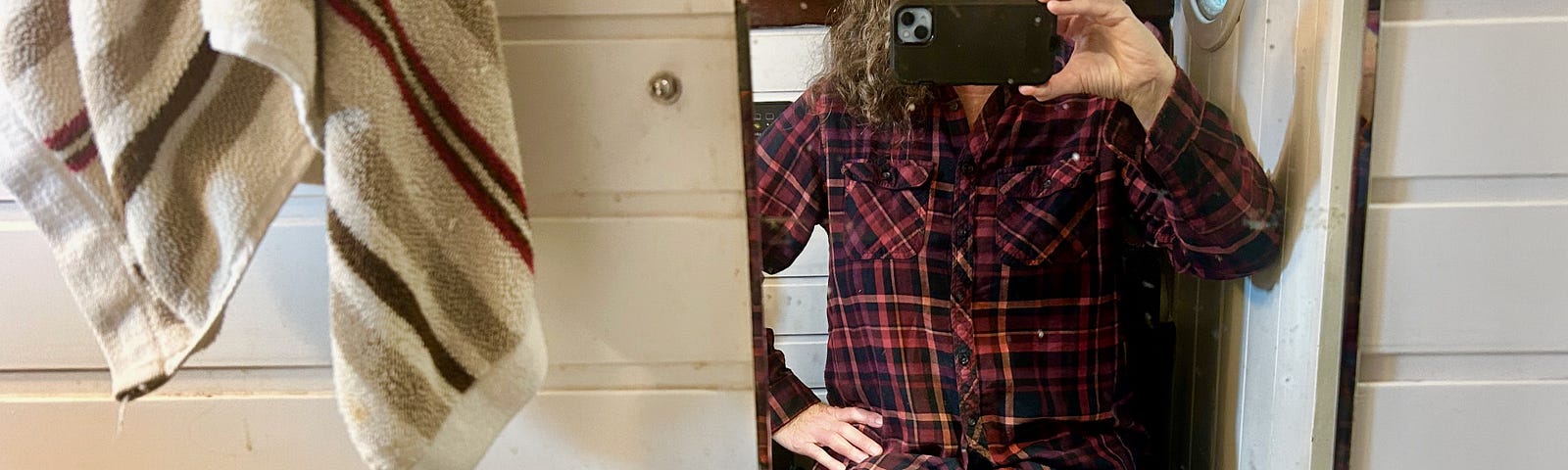 A woman sits on a toilet seat in a tiny bathroom. A mirror is in front of her as she takes a photo of herself. She has long grey hair and wears jeans and a plaid shirt.