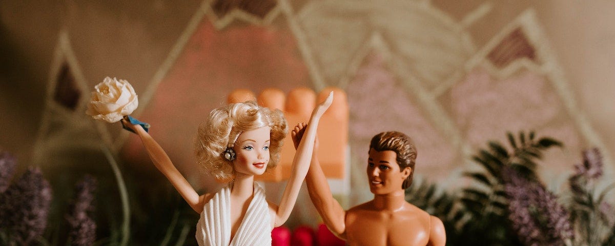 A Barbie doll wearing a white dress and holding a bouquet. She is standing beside a Ken doll wearing only board shorts.
