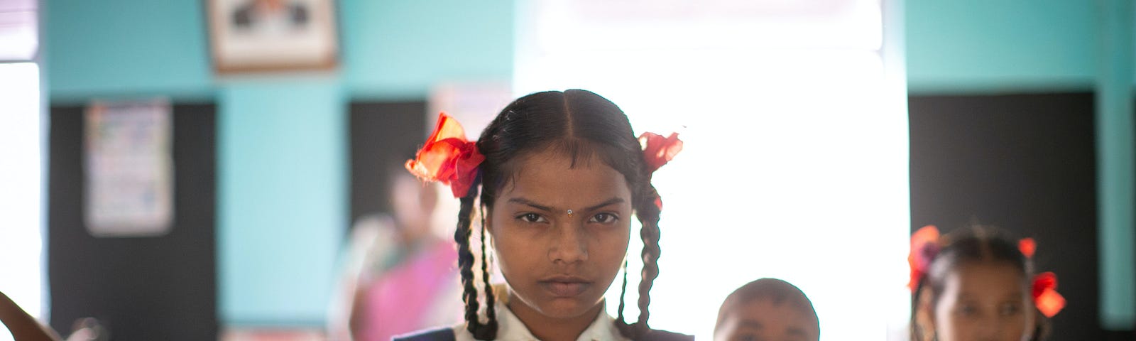 Picture of a girl in a school inform. Her hair is braided and she is looking intensely into the camera. In the background, there are five other children. Credit: Raj Rana via Unspalsh.
