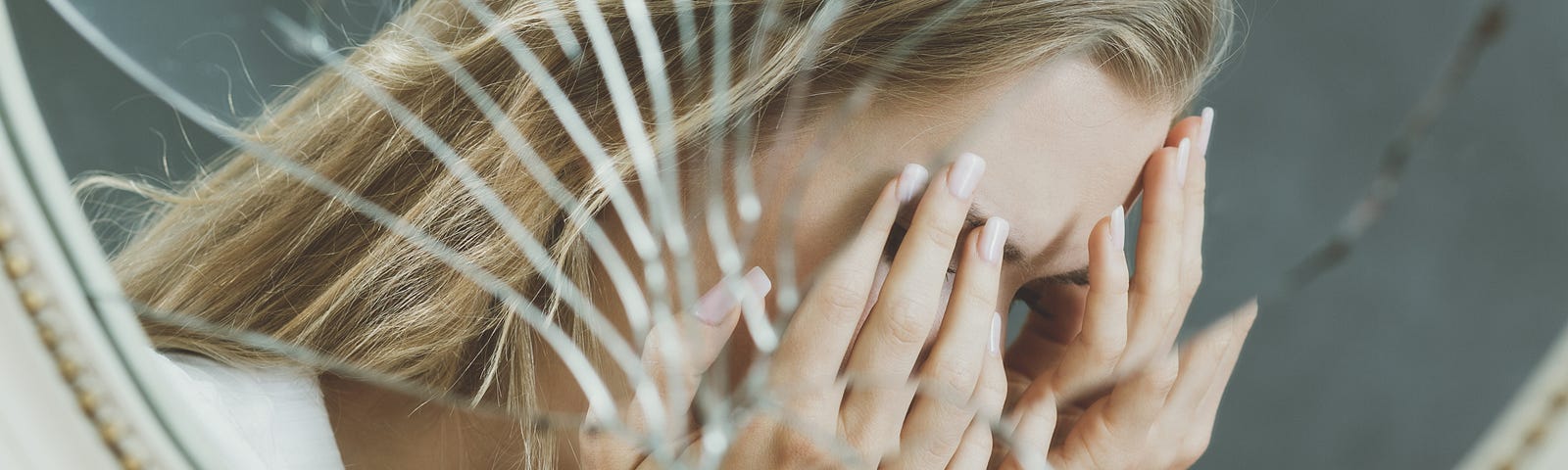 A woman holds her face in her hands and cries in the reflection of a shattered mirror.