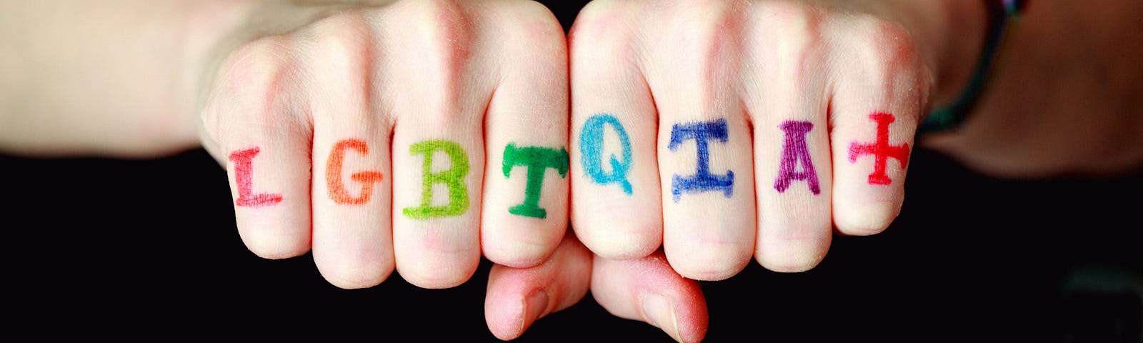 two fists pushed together. the letters LGBTQIA+ are painted in rainbow colors, one letter on each finger.