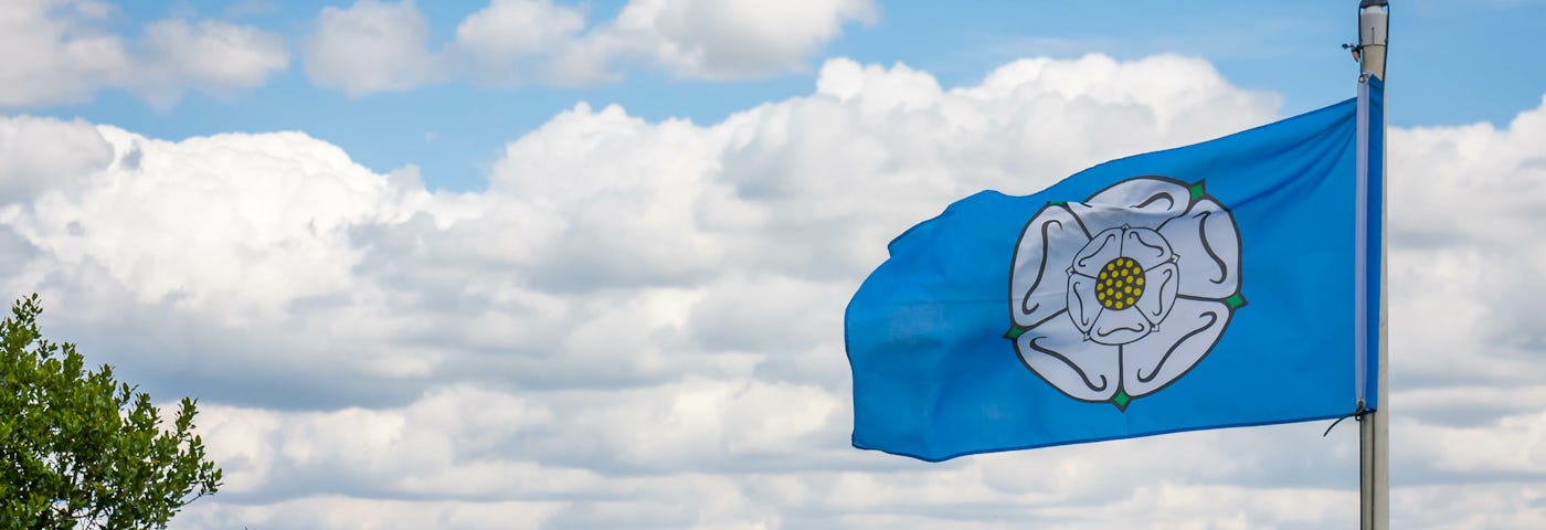 The Yorkshire Flag of a white rose on a blue background flies against a sunny sky over Yorkshire hills.