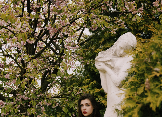 Young woman with thick black shoulder-length hair and red lipstick, wearing black under a beige rain-mac under a blossom tree next to a headstone.