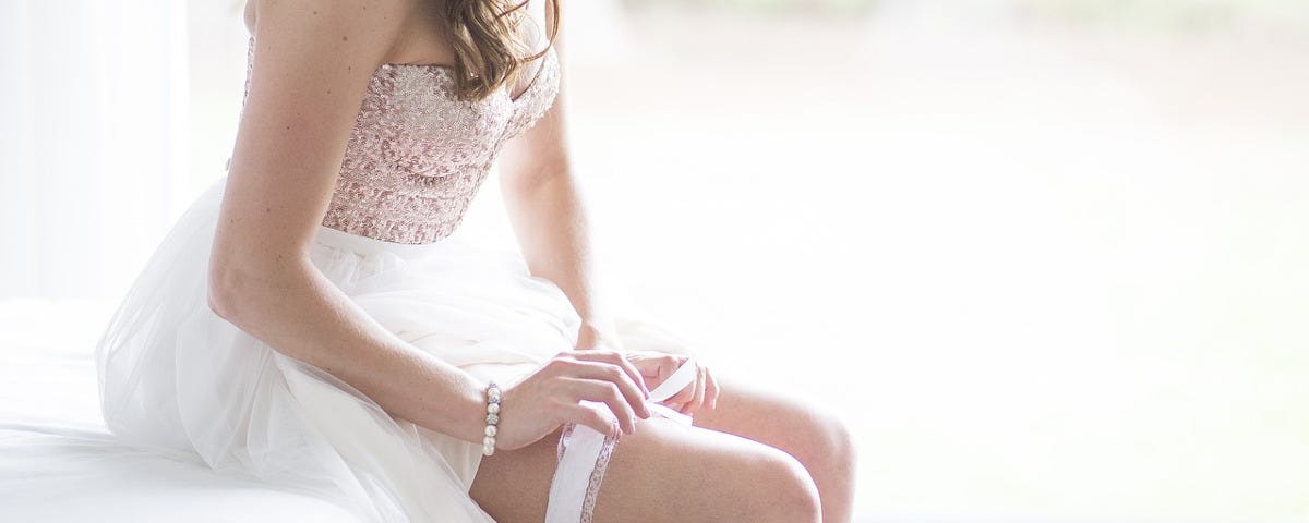 A bride, in a white dress, with white stockings, sitting on a white bed and pulling on a white garter.