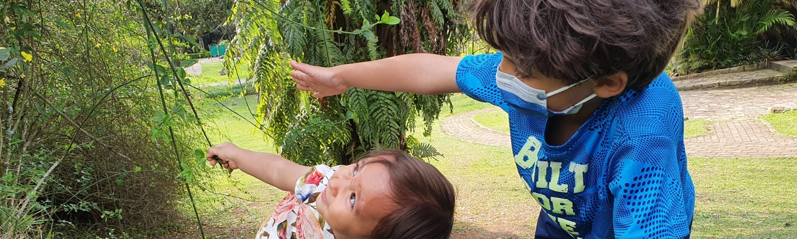 Foto de uma bebê e um menino escalando um morrinho verde. Ambos estão com uma mão no chão e outra erguida. A bebê sorri.