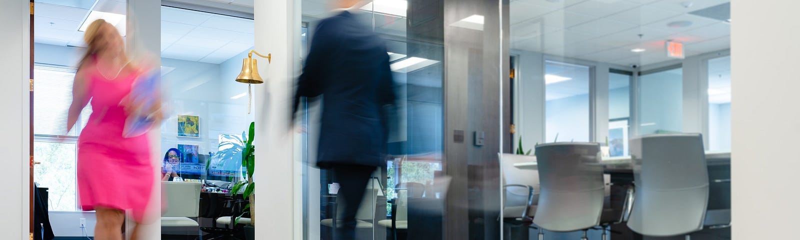 View of a office lobby with the backdrop of a conference room. A person in a pink dress and another in black suit are crossing each other. The faces are blurred.