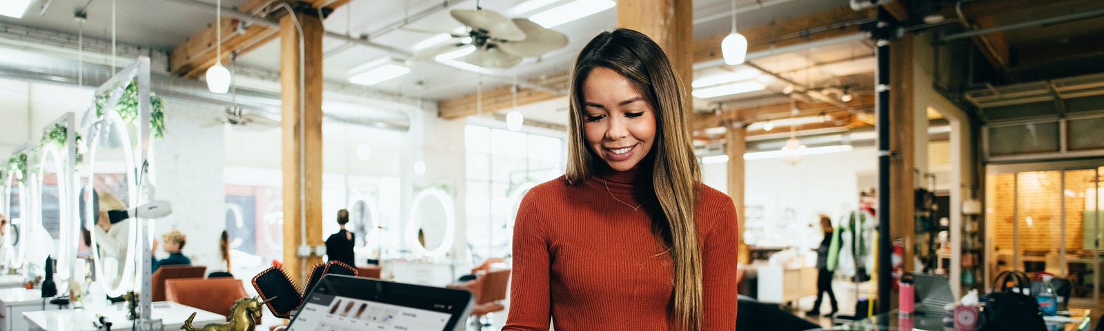 A woman with long hair swipes her credit card at a hair salon while someone behind the register operates a tablet device.