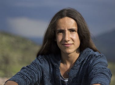 Photo of a young man with long black hair posed before a mountain range.