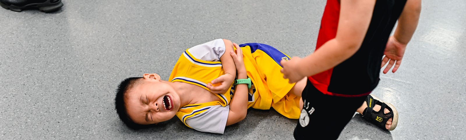 A small boy in a yellow sporting outfit clutching his stomach and crying. Another boy stands over him, dressed in a black and red kit.