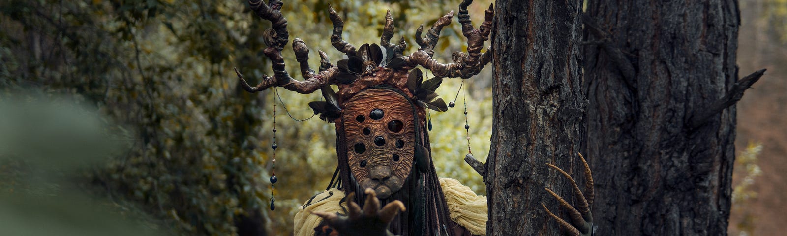 Monster with many eyes and horns stands next to tree and reaches with pointy fingers towards the camera.