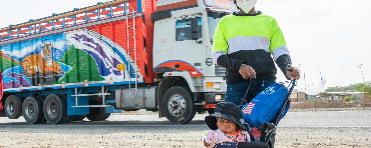 A woman wearing a baseball hat and a facemask stands with her child in a stroller in front of a truck.