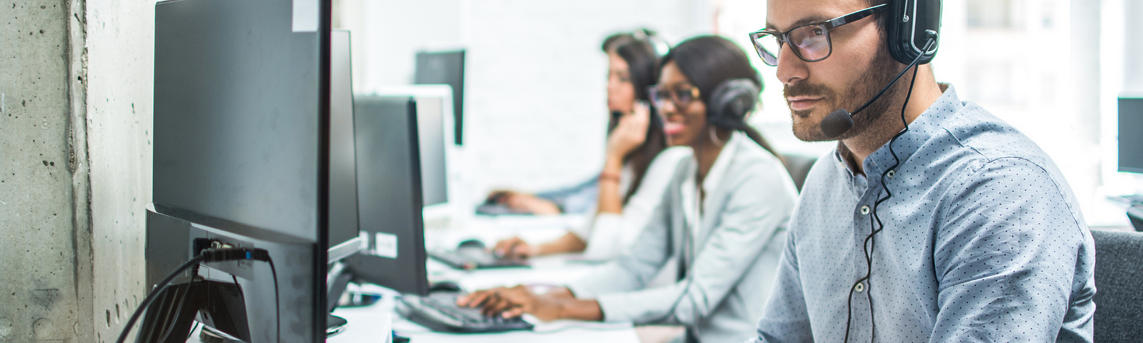 Three people wearing headsets sit in front of computers at an office. The man closest to the camera is in focus and has on glasses and a long sleeve shirt.