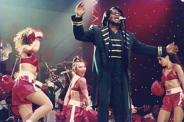 A photo of recording artist James Brown in concert with several female dancers in the background