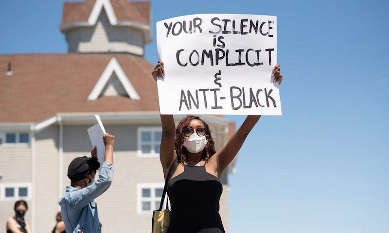 A protestor holds up a sign that reads “your silence is complicit and anti-black”