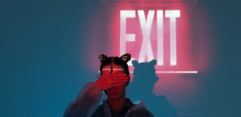 A woman holds her hands in front of her eyes as a bright red exit sign burns behind her.