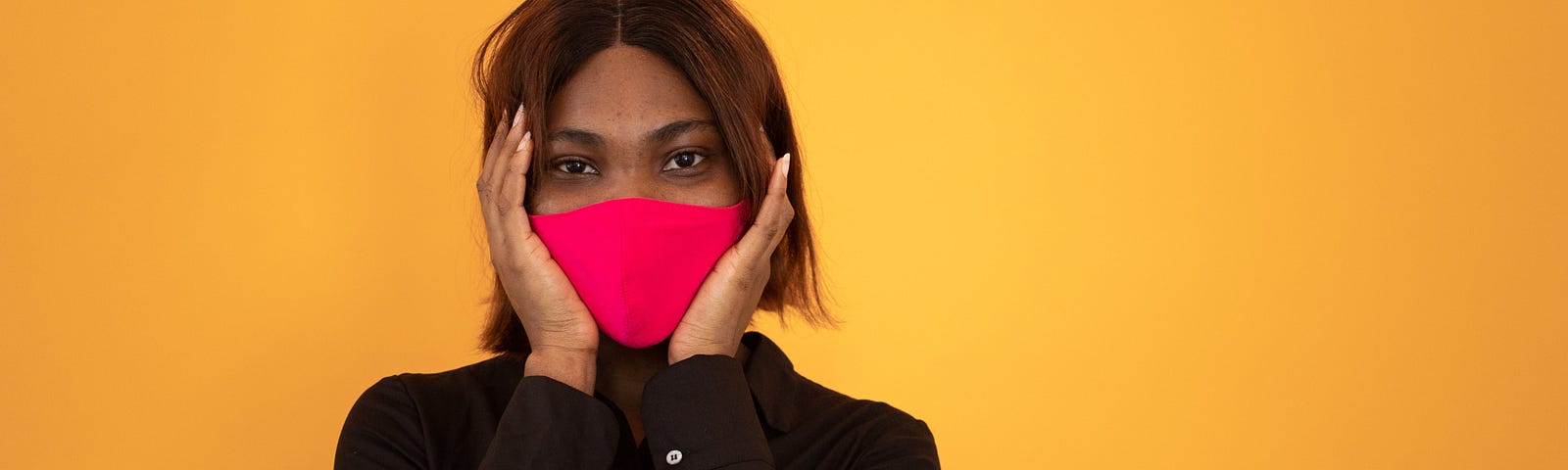 A woman wearing a long sleeved black button down shirt and a red face mask holds her hands to either side of her face and looks directly into the camera. She is posed against a dark yellow background.