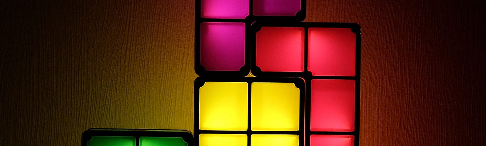 This is a tall rectangular picture with different shaped and colored blocks in front of a white wall. The background is dark but the shapes are internally lit. The shapes are from the traditional Nintendo Game — Tetris.