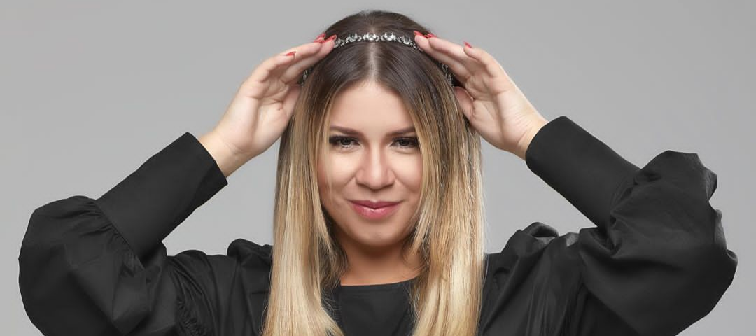 Marília Mendonça posa para foto com as mãos na tiara em cima da sua cabeça, veste uma blusa preta de mangas longas e bufantes.