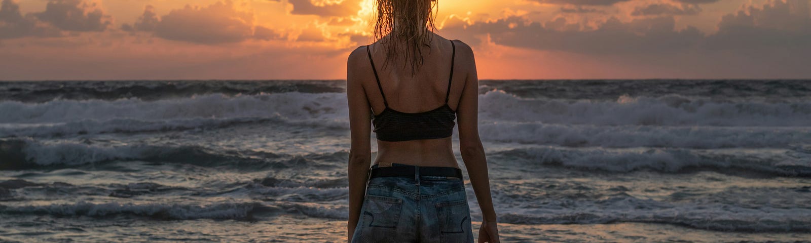 A woman in a black crop top and jean shorts watches the Sun set over the ocean on a beach.