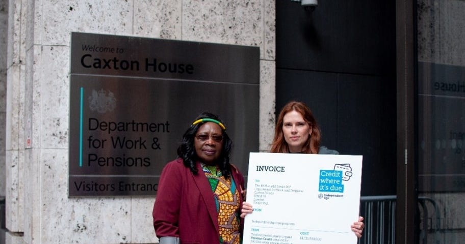 Photo of an older woman wearing a colourful dress and using a crutch, stood next to a younger woman who holds a large invoice prop telling Mel Stride that the Government’s Pension Credit payments are overdue.