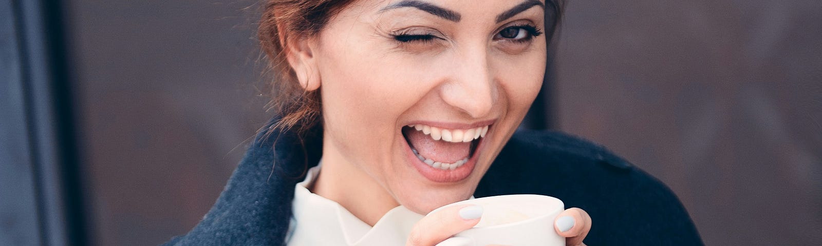 woman winking at camera, holding coffee cup and smiling — brown hair, white blouse, blue coat thrown open wide