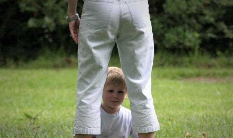 Three year old boy seated in the grass between his mother’s legs.