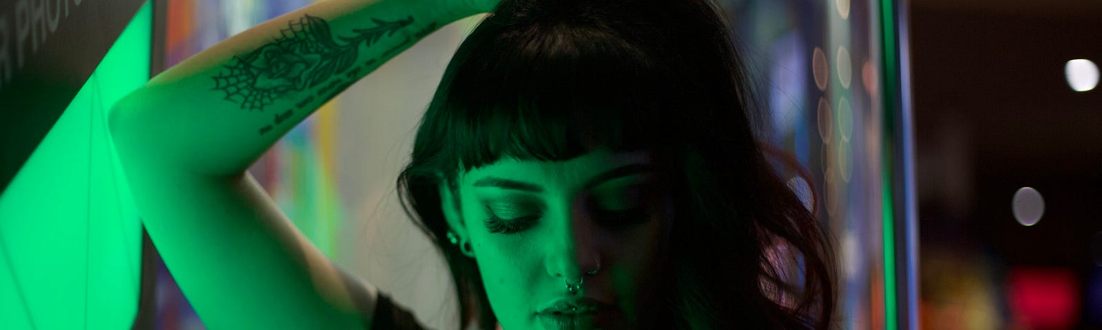 A young woman with dark hair and tattoos is standing in an arcade and painted green with light.