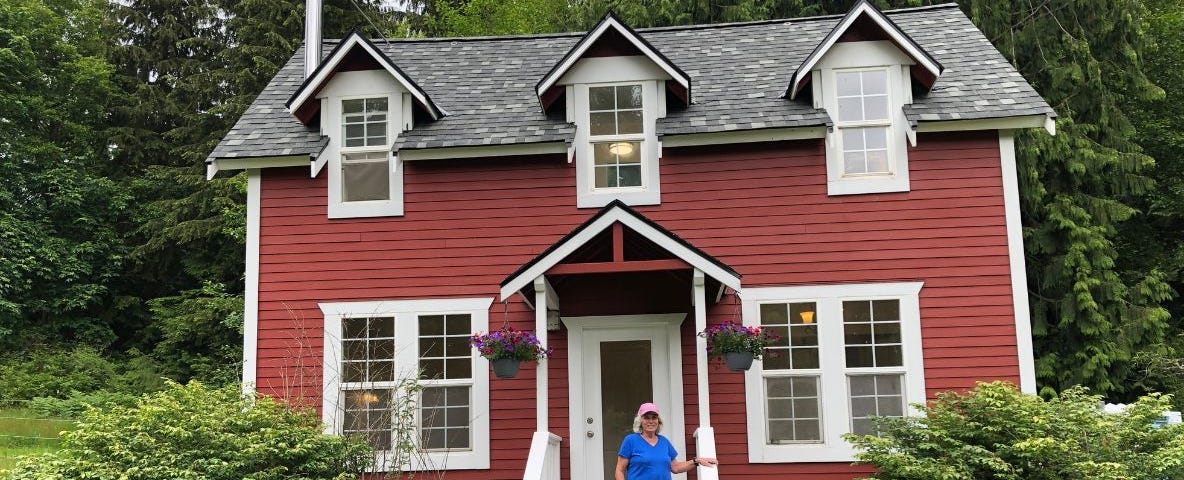 A 70 year old woman on the steps of her farmhouse-style red cottage near the woods.