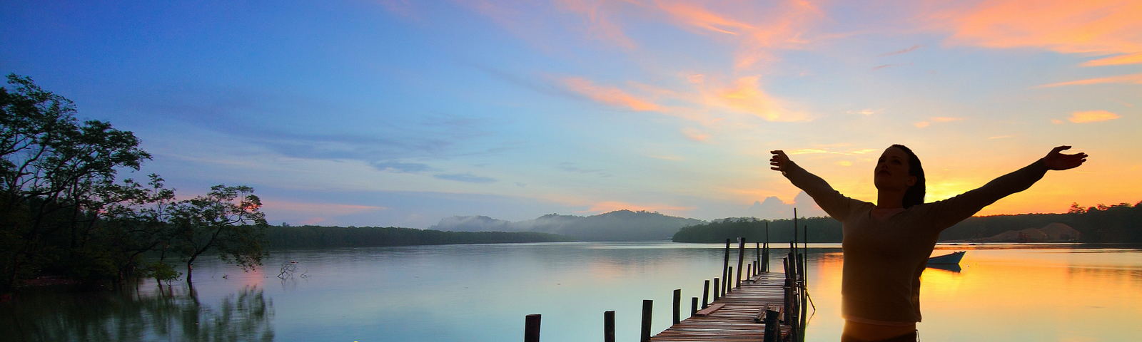 woman standing on dock, arms raised, looking out at the sun setting in the distance. Words in lower left corner: see the world with new eyes