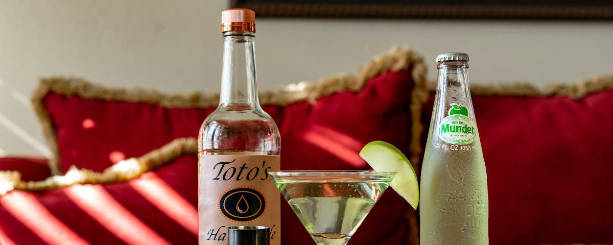 A cocktail in a martini glass, garnished with a thin slice of apple. The glass is on a coffee table in a living room, and surrounded by a bottle of spirits, a smaller bottle of a green soft drink, and a shot measure.