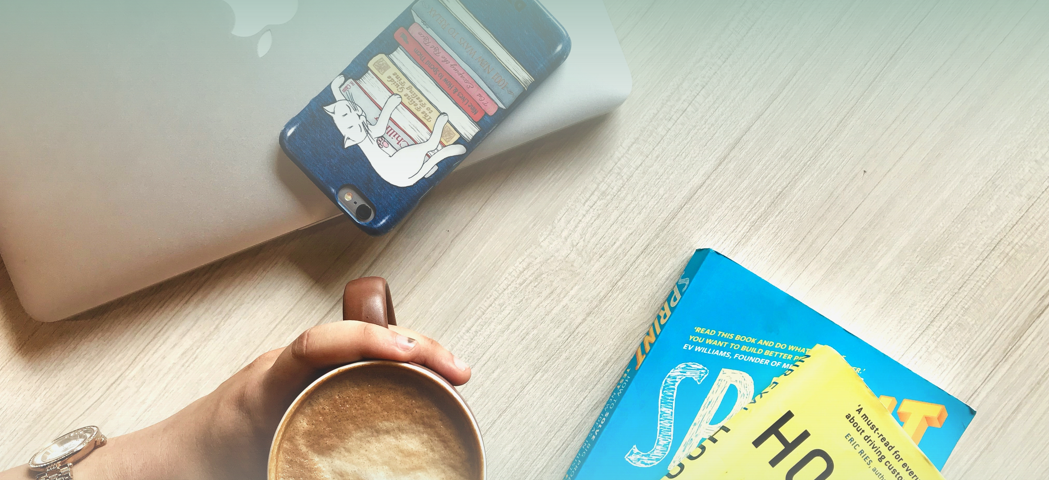 A person holding a coffee while at their desk. On the desk is a smart phone and two business books.