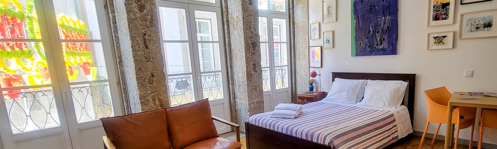 The inside of a studio apartment in Braga with granite walls, three French doors with windows, a small loveseat, a bed and a table and chair.