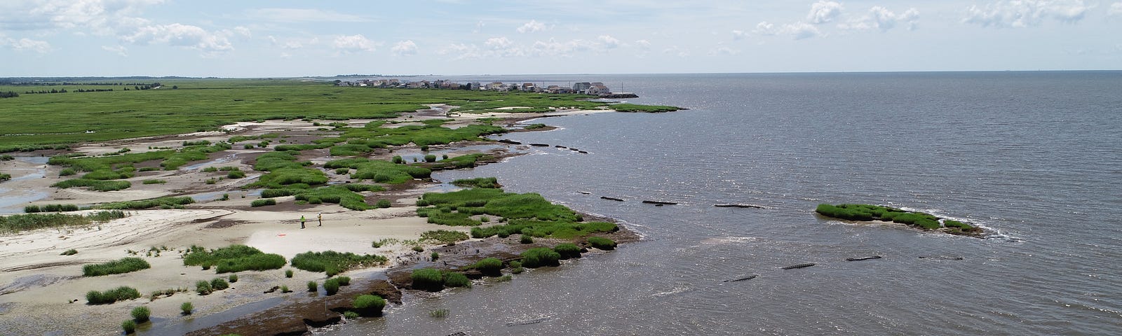 This aerial view shows the coastline with beach area and green marsh to the left. The ocean is to the right. In the water just off the sloping shoreline are breakwaters. There’s distance between each breakwater. A small peninsula no longer visibly connected to the coastline is visible. There is a row of residential structures along the water in the background.