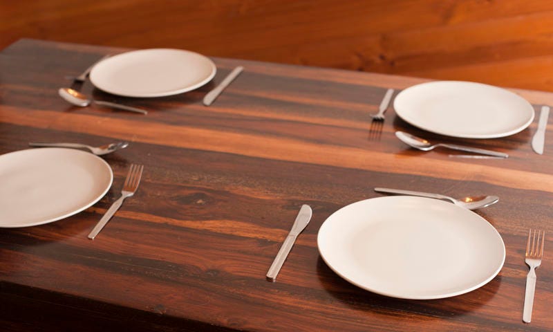 Four basic place settings on a wooden dinner table with plates and cutlery but no linen or placemats, closeup view